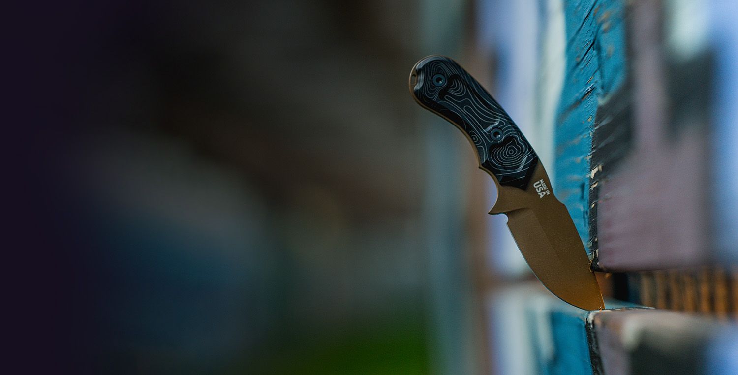 Bronze-bladed HAViK karambit knife with a topographic handle resting against a blue-painted wood fence