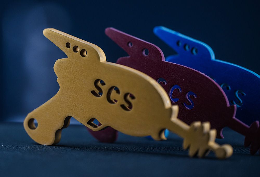 Three anodized aluminum ray-gun cutouts in yellow, red, and blue with SCS lettering