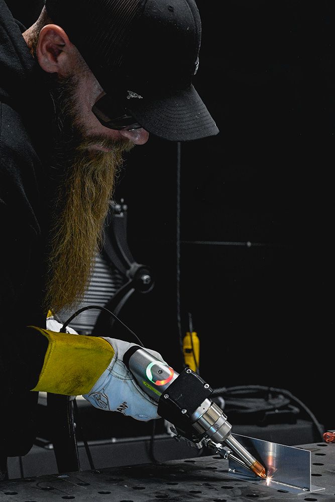 Bearded welder using a handheld laser welder on a perforated metal workbench