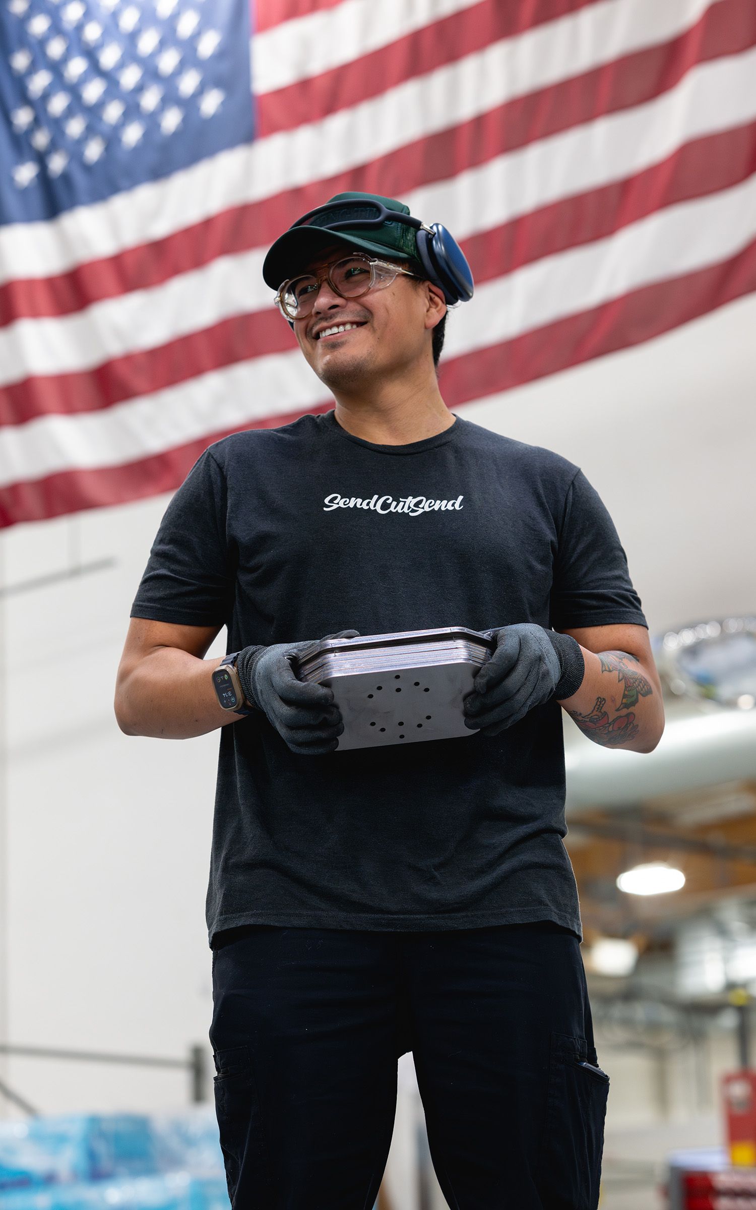 Worker in hard hat standing beside a large American flag inside a steel fabrication facility