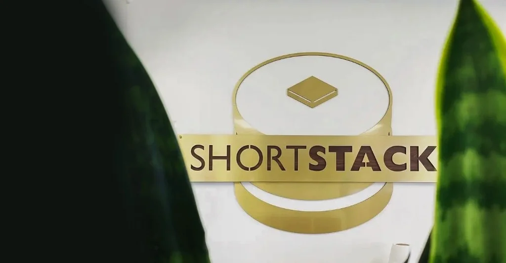 ShortStack office sign in gold lettering with a stacked-disc logo, framed by out-of-focus greenery